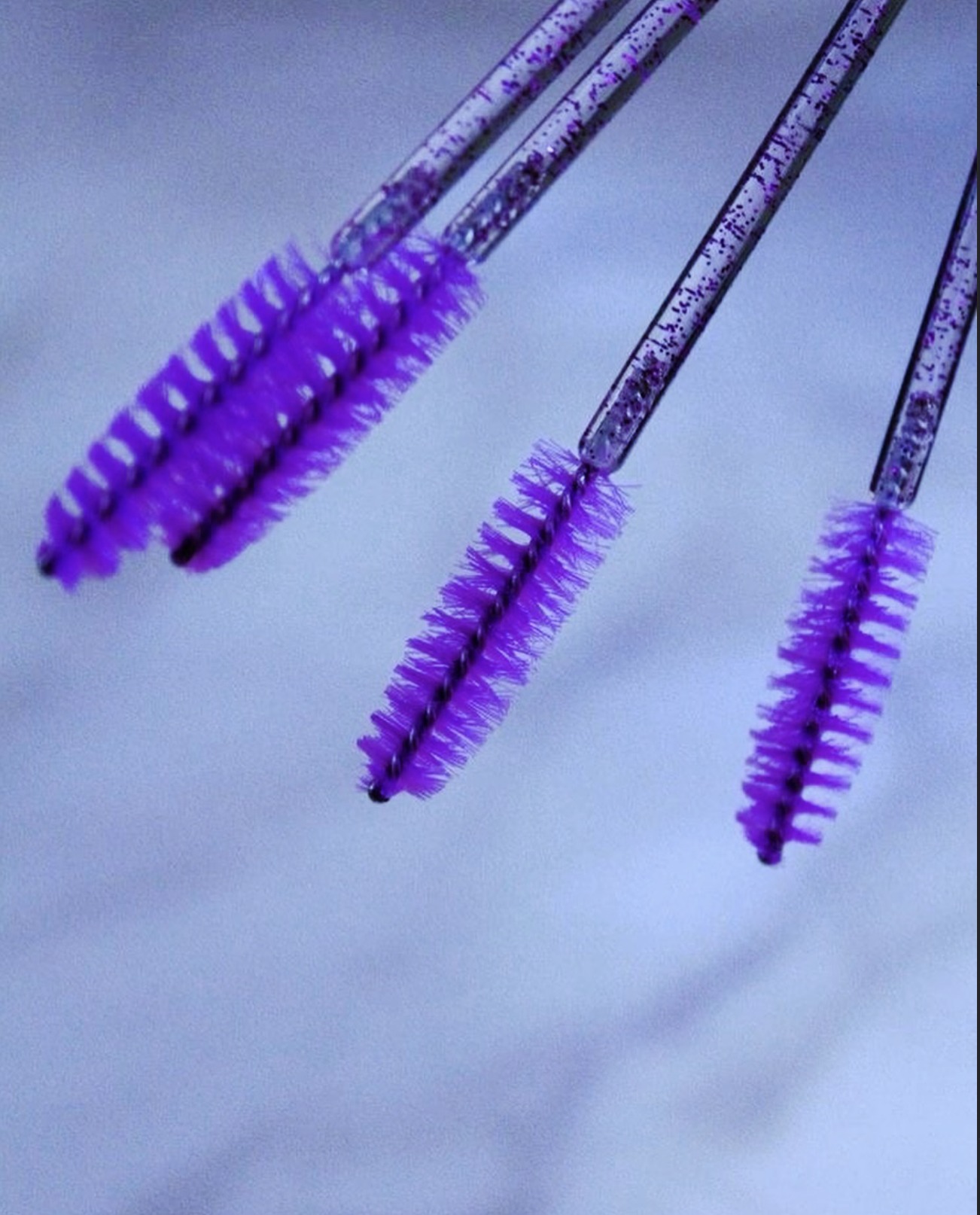 Purple glitter mascara wands on a soft blue backdrop — Lashes by Dee studio tools